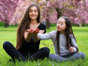 Big sister and little sister sit together on a lawn with cherry blossoms behind them