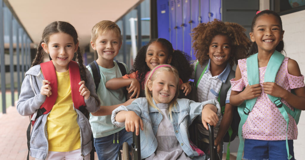 Front view  of happy school kids standing in  outside corridor at school while a Caucasian schoolgirl is sitting on wheelchair in foreground